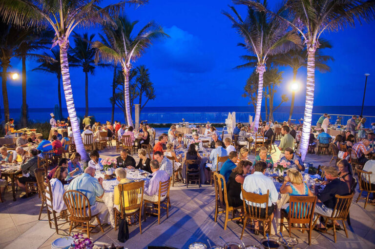 people seated a dinner tables under palm trees at an outdoor evening event