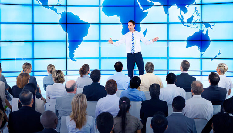 man presenting on stage at a corporate meeting event with a world map stage backdrop